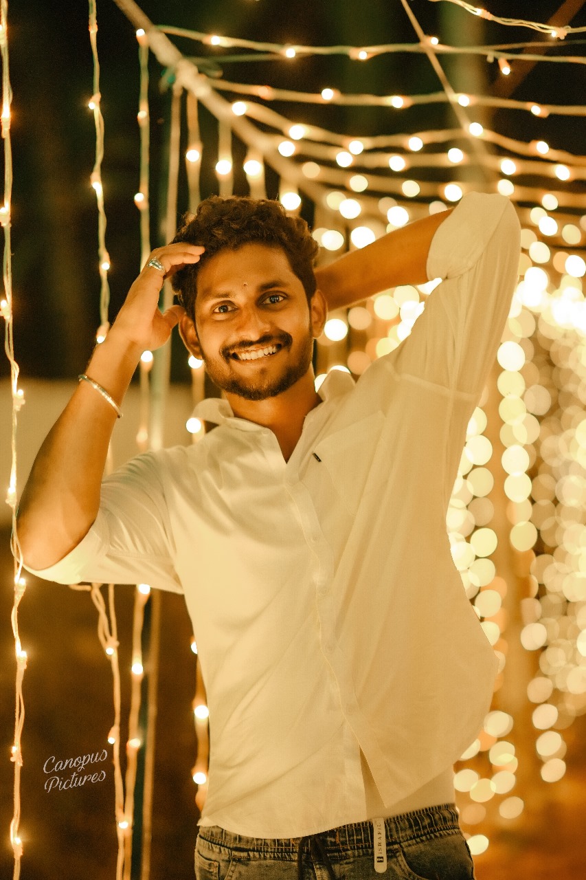 Cheerful software developer in Kochi, Kerala, wearing a white shirt and
						denim, posing under festive string lights, radiating positivity.