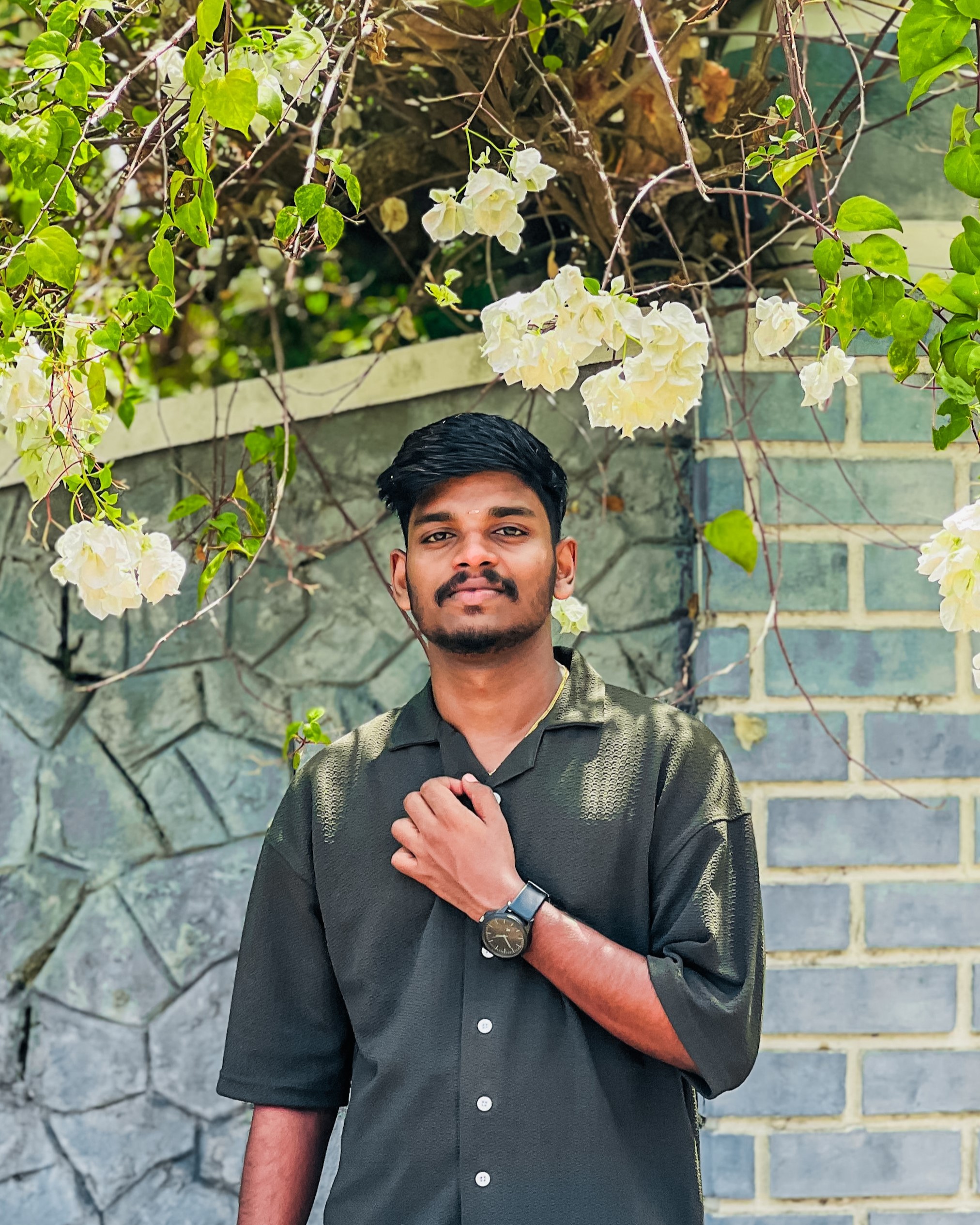 Portrait of a young software developer in Kochi,
						Kerala, standing confidently under blooming white flowers, wearing a dark green shirt