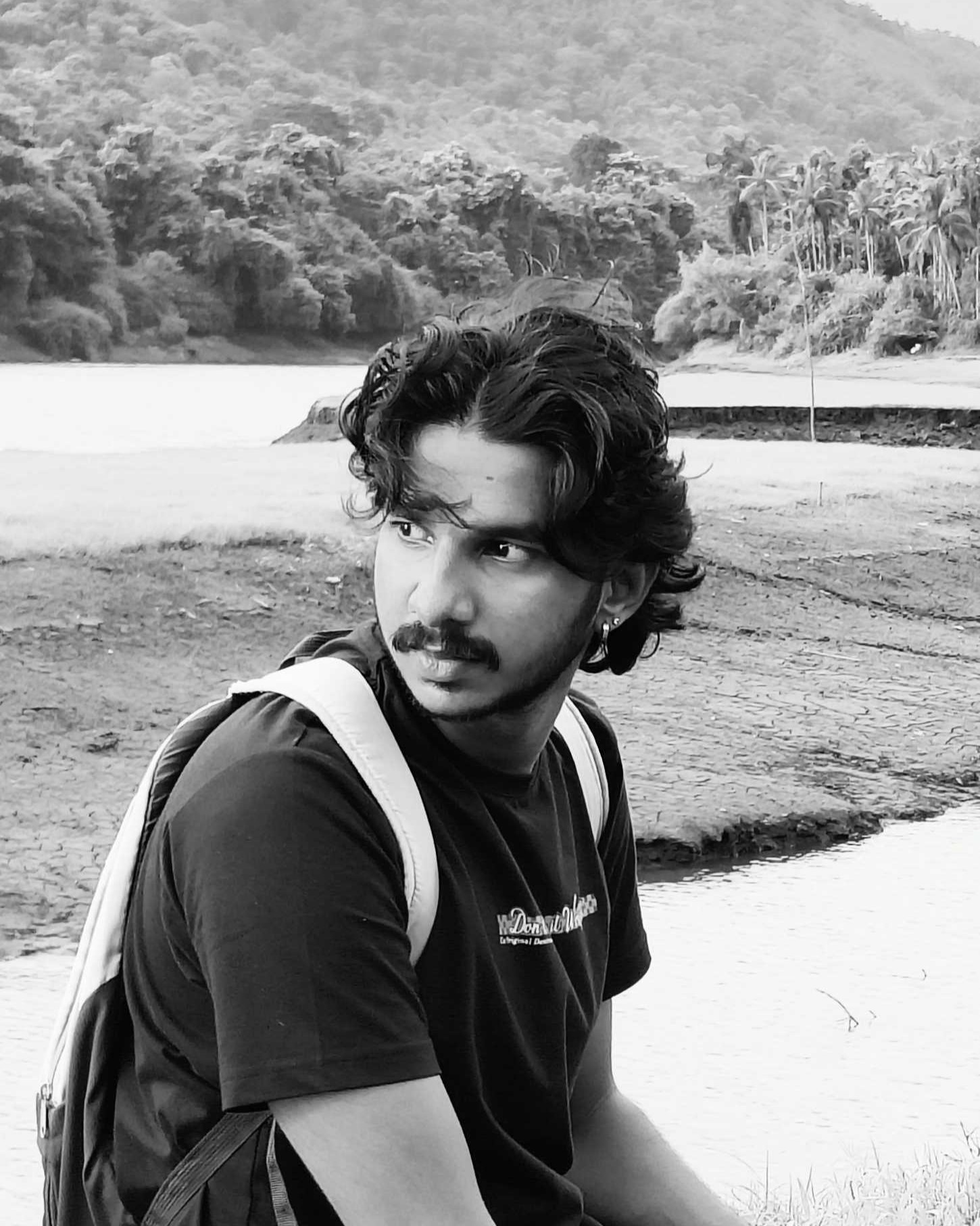 Monochrome image of a young software developer in Kochi, Kerala,
						seated outdoors with a serene lake and forest in the background, wearing a casual black t-shirt
						and backpack.