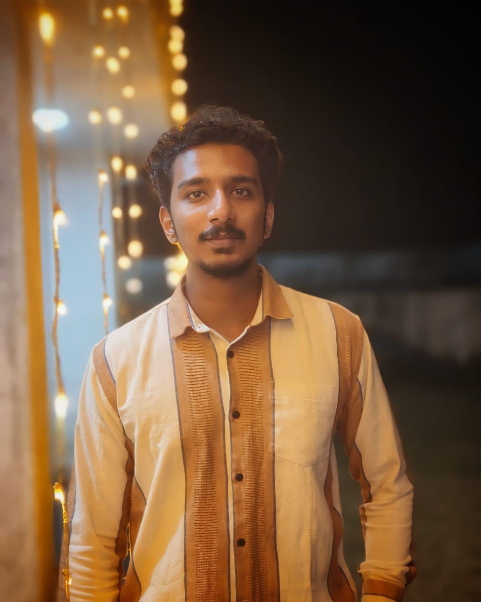 Young software developer in Kochi, Kerala, dressed in a light beige shirt,
						standing against a festive backdrop illuminated with warm lights, radiating a cheerful
						expression.