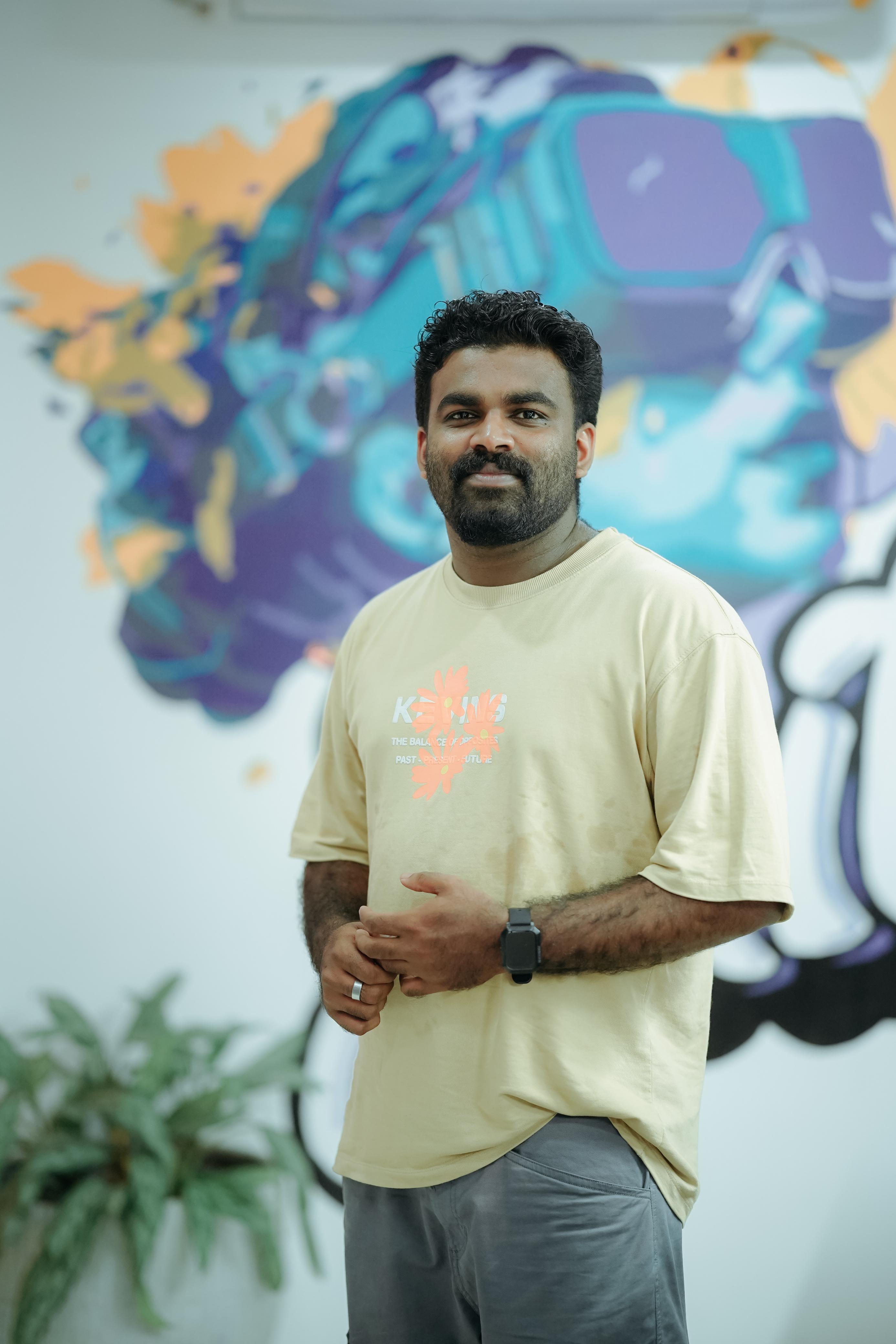 Creative software developer in Kochi, Kerala, wearing a casual beige
						t-shirt, standing confidently against a vibrant artistic mural background.
