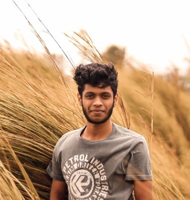 Outdoor portrait of a software developer in Kochi, Kerala,
						standing amidst tall grass, wearing a casual grey t-shirt with a rustic and natural backdrop.