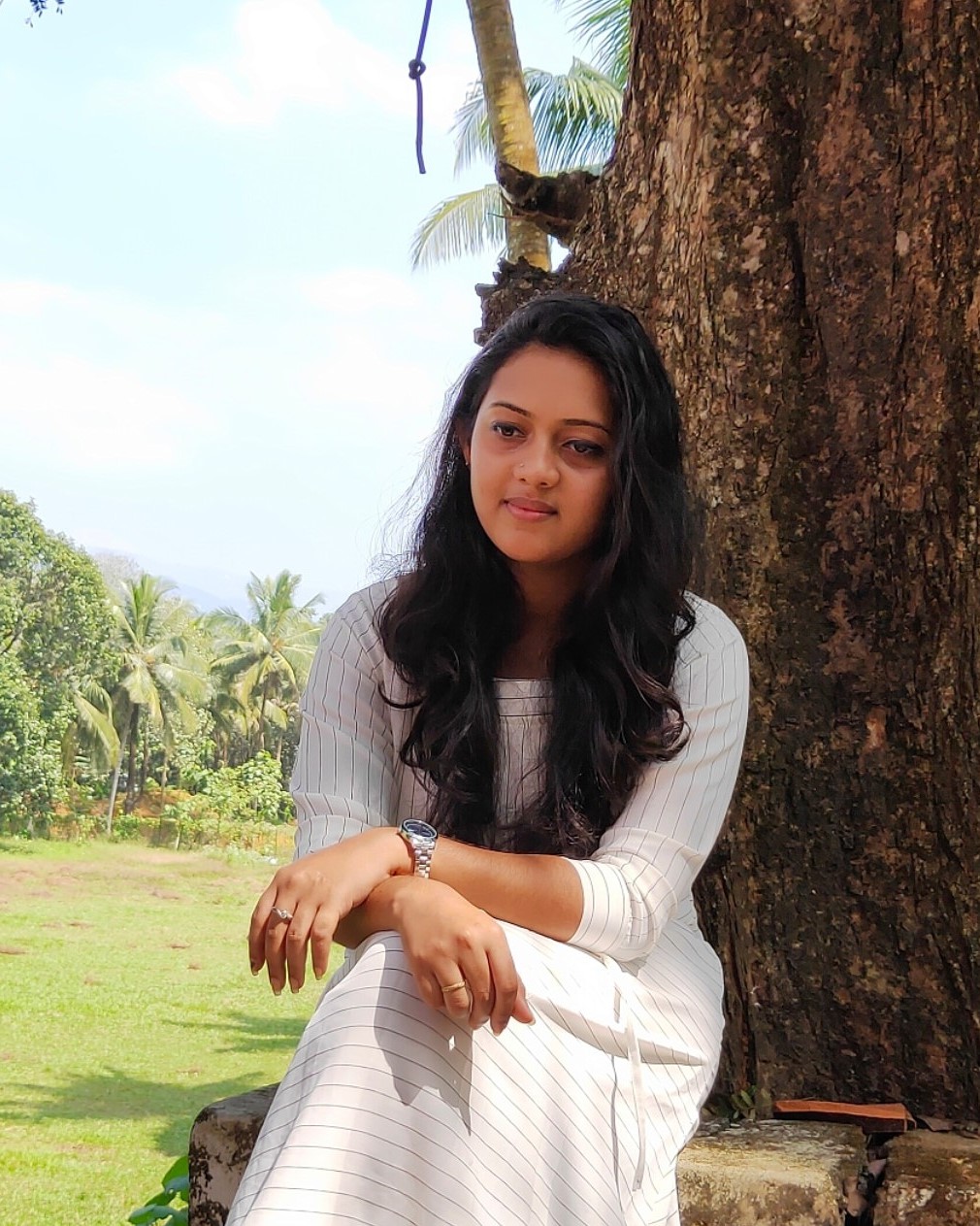Thoughtful software developer in Kochi, Kerala, seated
						under a tree, wearing a white-striped dress, with lush greenery in the background.