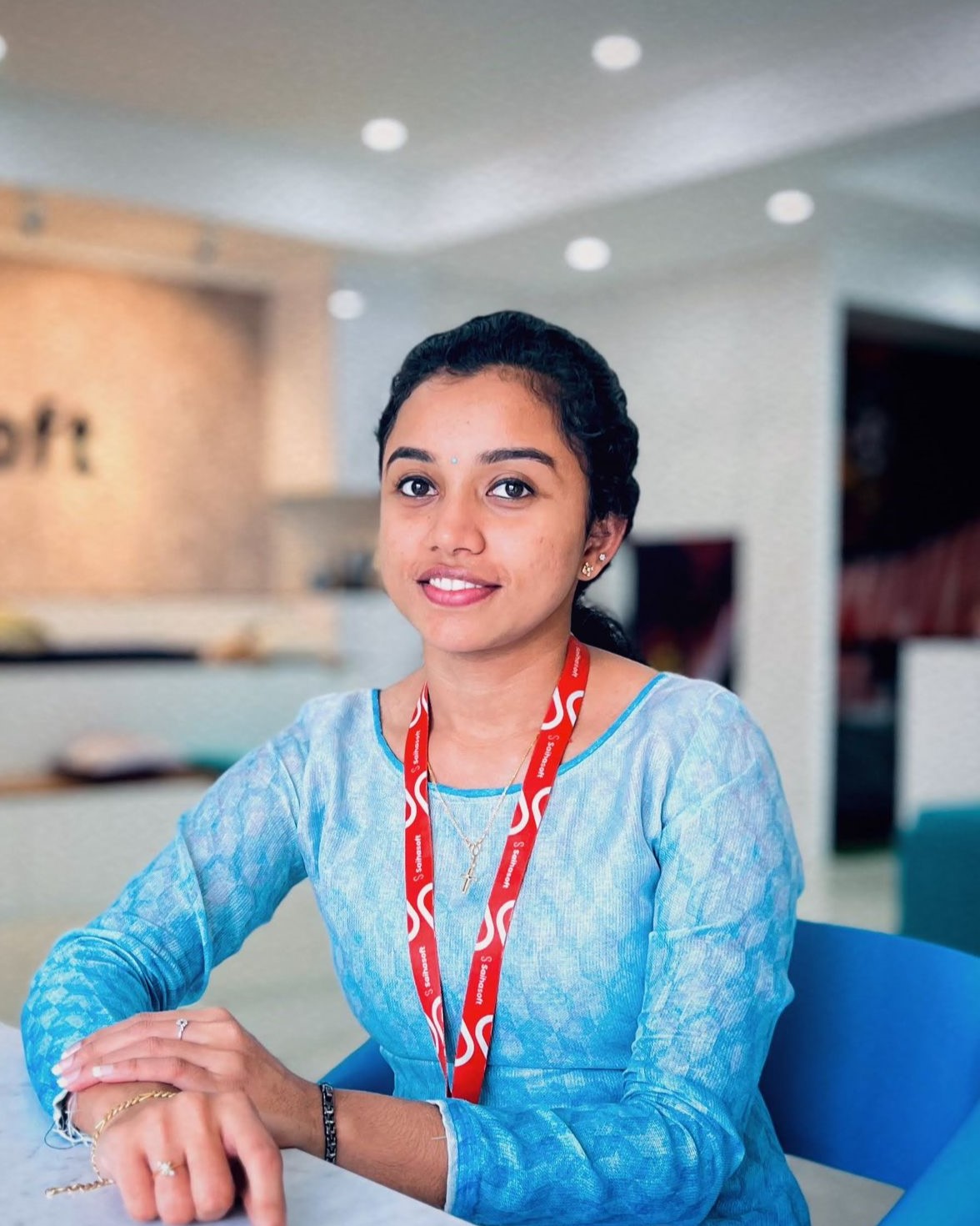 Professional software developer in Kochi, Kerala, dressed in a
						traditional outfit, seated at a desk in a modern office space with a warm smile.