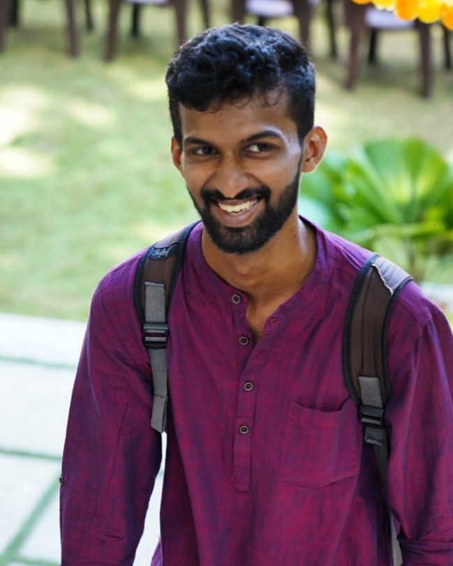 Smiling software developer in Kochi, Kerala, wearing a
						traditional purple kurta with a backpack, captured in a lively outdoor setting.