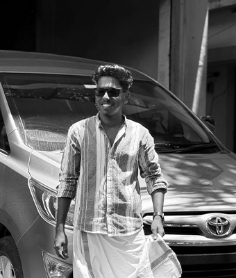 A young man is captured in a black and white photo standing in front of an SUV. He is wearing sunglasses, a striped shirt, and a traditional white dhoti, holding one corner. He is looking off to the side with a casual, confident smile.