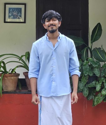 Young software developer in Kochi, Kerala, wearing a light blue shirt and a traditional white mundu, standing in front of a heritage Kerala house with potted plants.