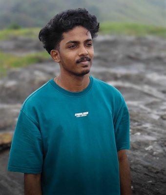 Young software developer in Kerala with curly hair, wearing a teal t-shirt, standing on a rocky hilltop with misty green mountains behind him.