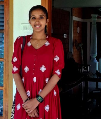 Cheerful young professional woman in Kerala, wearing a red dress with white embroidered motifs, standing at the doorway of a traditional house.