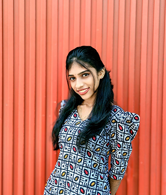 Young software developer in Kerala, dressed in a geometric-patterned outfit, standing against a vibrant red background, exuding a cheerful and confident presence.