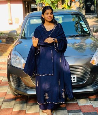 Young professional woman in Kerala, dressed in a navy blue embroidered churidar with a matching shawl, standing in front of a grey car on a tiled driveway.