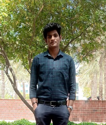 Young software developer wearing a dark checked shirt and grey trousers, standing confidently under a leafy tree with palm trees in the background.