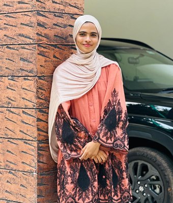 Young professional woman in Kerala wearing a beige hijab and a peach embroidered top, leaning against a textured brick pillar beside a black car.