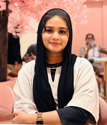 Smiling young woman in Kerala wearing a black scarf and a cream top, seated in a cafe under a pink cherry blossom tree.