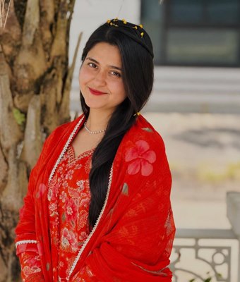 Young professional woman in Kerala with long dark hair, wearing a red floral churidar with a matching red dupatta, smiling beside a tree outdoors.