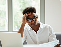 A man wearing glasses is seated at a desk, working on a laptop surrounded by papers.